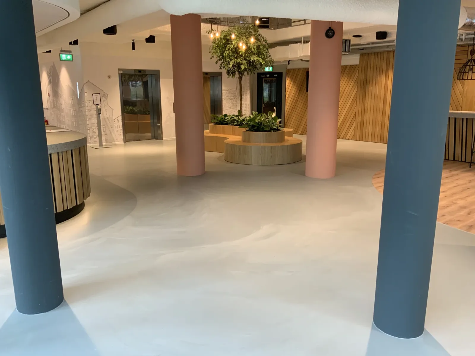 Lichtgrijze Comforpur 635 Betonlook gietvloer in een ruime ontvangsthal met ronde zitbanken, beplanting en liften